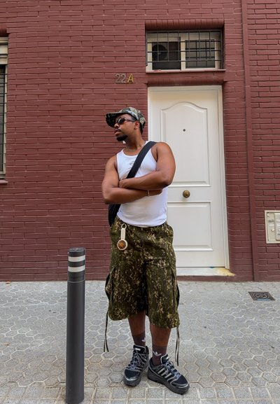 Hombre con gafas de sol, camiseta blanca sin mangas, pantalones cortos de camuflaje y gorra, de pie con los brazos cruzados en la acera frente a la puerta de un edificio de ladrillo rojo.