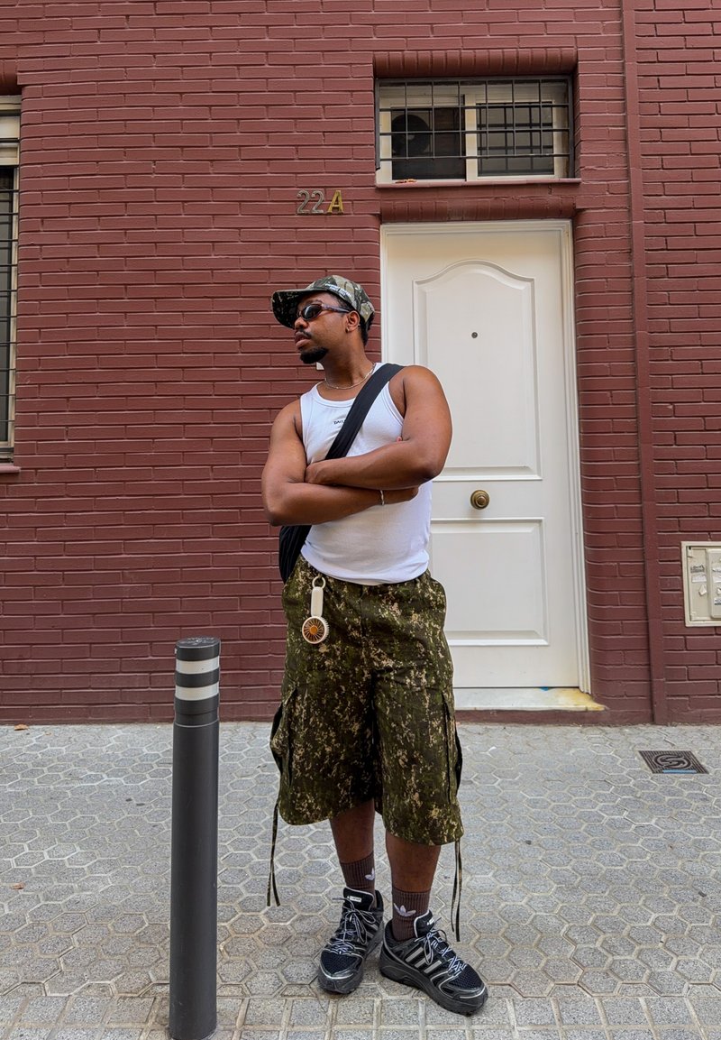 Homme portant des lunettes de soleil, un débardeur blanc, un short camouflage et une casquette, debout les bras croisés sur le trottoir devant la porte d’un bâtiment en briques rouges.