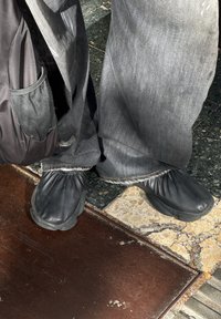 Chaussures noires en cuir souple avec une partie supérieure élastique froncée ; portées avec un pantalon en denim foncé et ample. L'arrière-plan comprend un pavé texturé.