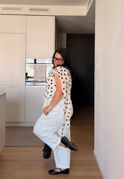 Mujer con pantalones blancos y blusa negra con lunares posando dentro de casa, sosteniendo un zapato negro mientras lleva el otro puesto, de pie sobre un suelo de madera.