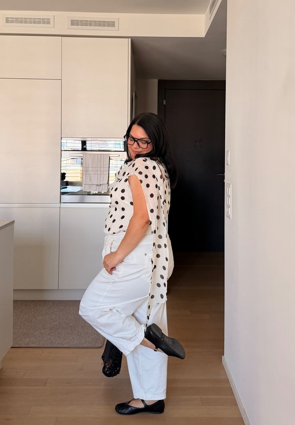 Vrouw in witte broek en zwarte blouse met stippen poseert binnen, houdt één zwarte schoen vast terwijl ze de andere draagt, staand op een houten vloer.