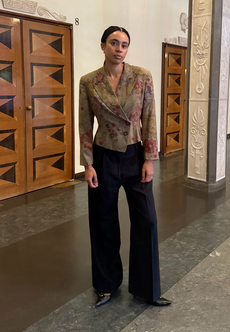 Femme debout à l'intérieur portant un blazer à motif floral, un pantalon noir à jambes larges, et des chaussures noires pointues dans un couloir décoré.