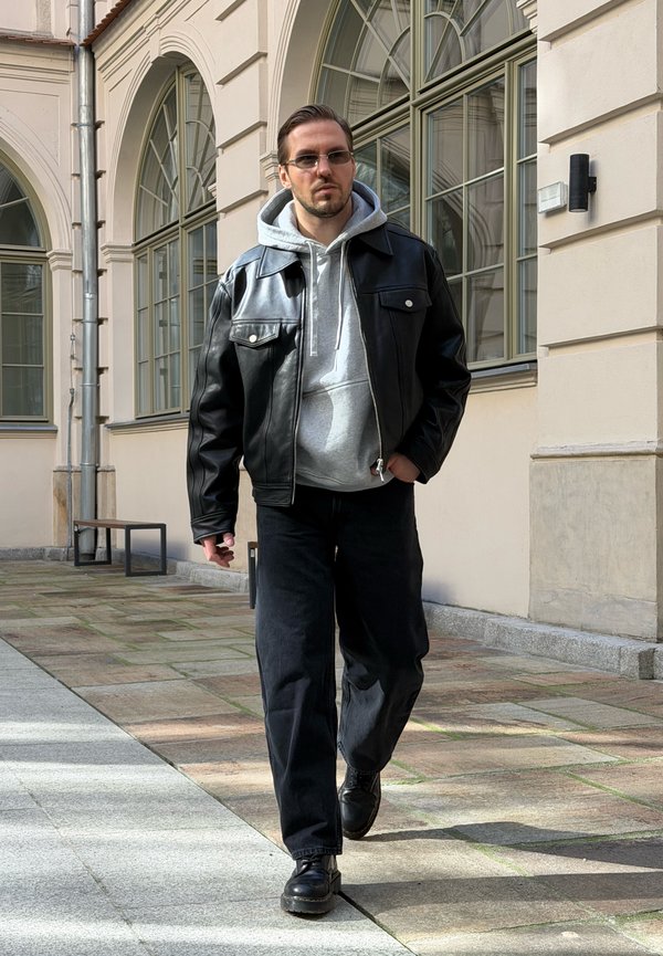 Hombre con chaqueta de cuero negra, sudadera con capucha gris, pantalones negros y gafas de sol caminando afuera cerca de ventanas arqueadas y un edificio beige.