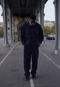 Mörk denimjacka och matchande byxor, med knäppning framtill och bröstfickor. Bärs med svarta skor. Urban miljö under en bro.