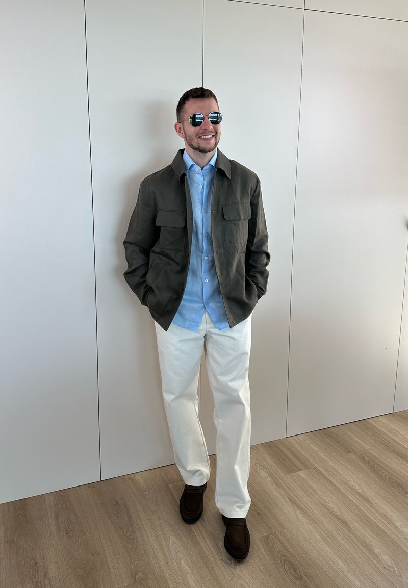 Hombre con gafas de sol, chaqueta oscura, camisa azul clara, pantalones blancos y zapatos oscuros, de pie sobre un suelo de madera clara frente a una pared blanca.