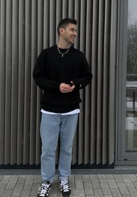 Homme portant un pull noir, un jean bleu clair et des baskets noires, debout et souriant contre un mur gris texturé près d'une porte en verre.
