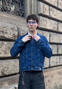 Kék, hosszú ujjú farmerdzseki, patentos elülső része, mellzsebekkel. Fekete farmernadrág, kulcstartóval rögzítve. Világos színű fal háttér.