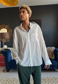 Jeune personne aux cheveux blonds bouclés portant une chemise blanche ample boutonnée et un pantalon vert, se tenant à l'intérieur près d'un canapé bleu et d'une lampe.