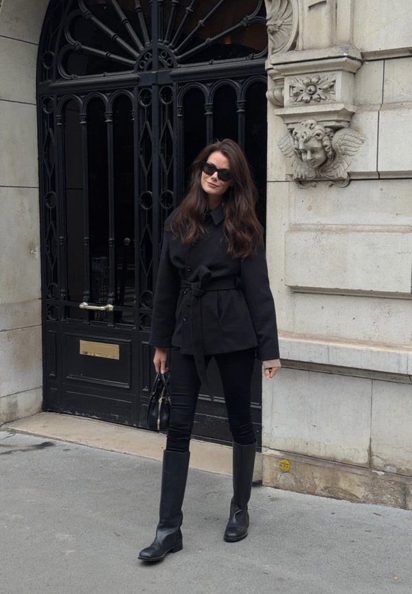 Una mujer con abrigo negro, pantalones, gafas de sol y botas está frente a un edificio de piedra ornamentado y lleva un bolso pequeño.