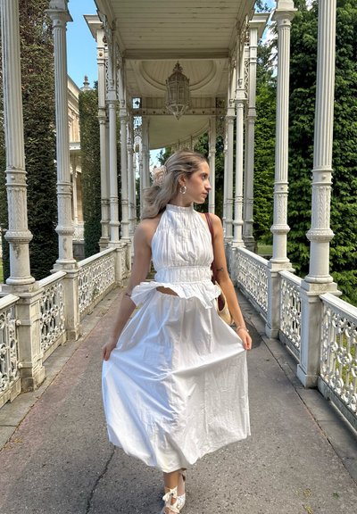 Top blanco con cuello halter, con textura plisada y volantes en capas, combinado con una falda larga y fluida blanca, situado frente a un hermoso arco blanco.