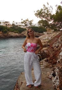 Vrouw in zonnebril, gekleed in een roze bloemen top en witte broek, staat op een rotsachtige waterkant met trappen en Mediterraan huizen op de achtergrond.