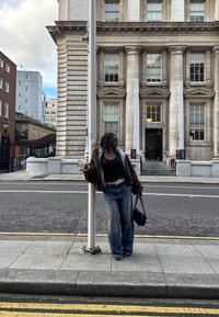 Femme en jean et veste sombre appuyée contre un réverbère sur le trottoir, tenant un verre et un sac, avec un bâtiment classique en pierre en arrière-plan.