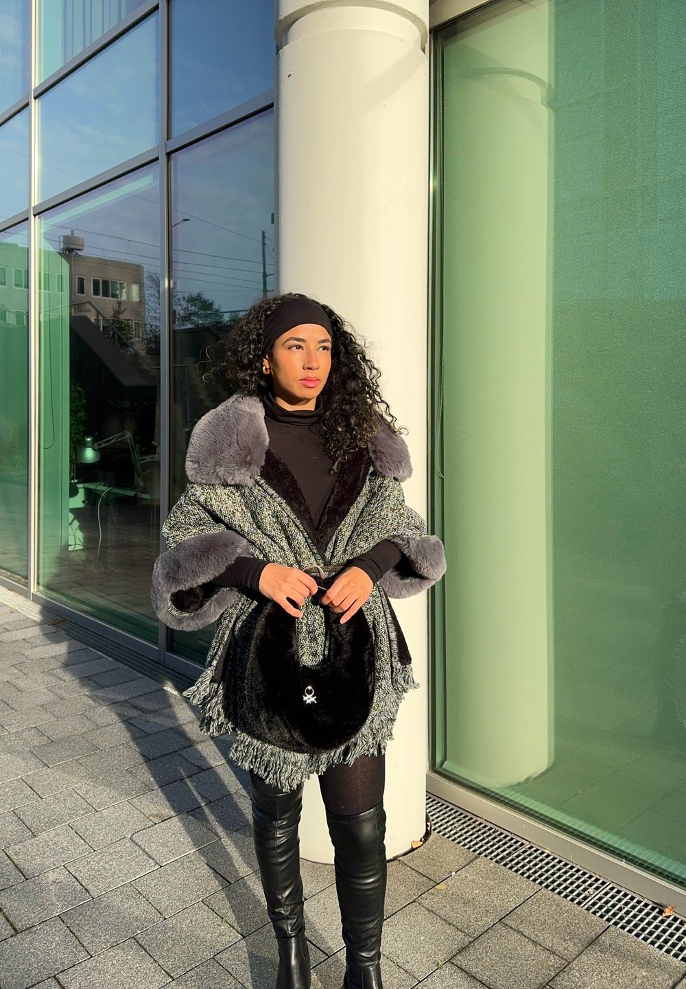 Šedý a čierny textúrovaný kabát s umelou kožušinou na golieri, cez čierny outfit. Drží čiernu tašku s strieborným emblémom. Osvetlené mestské pozadie.