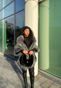 Šedý a čierny textúrovaný kabát s umelou kožušinou na golieri, cez čierny outfit. Drží čiernu tašku s strieborným emblémom. Osvetlené mestské pozadie.