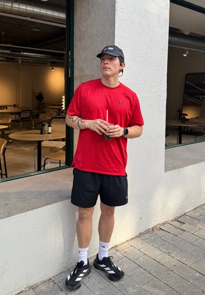 Camiseta atlética roja de manga corta con un logo, pantalones cortos negros y zapatillas negras. Sujetando una bebida con una pajita, de pie junto a una ventana.