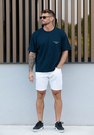 Camiseta de algodón azul marino de manga corta con un logotipo impreso, combinada con pantalones cortos de mezclilla blancos y zapatillas negras. Modelo lleva gafas de sol.