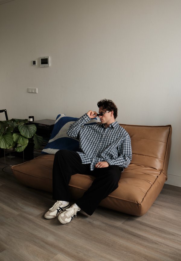 Uomo con occhiali da sole e camicia a quadri seduto su un divano basso in pelle marrone in una stanza minimalista con pavimento in legno.