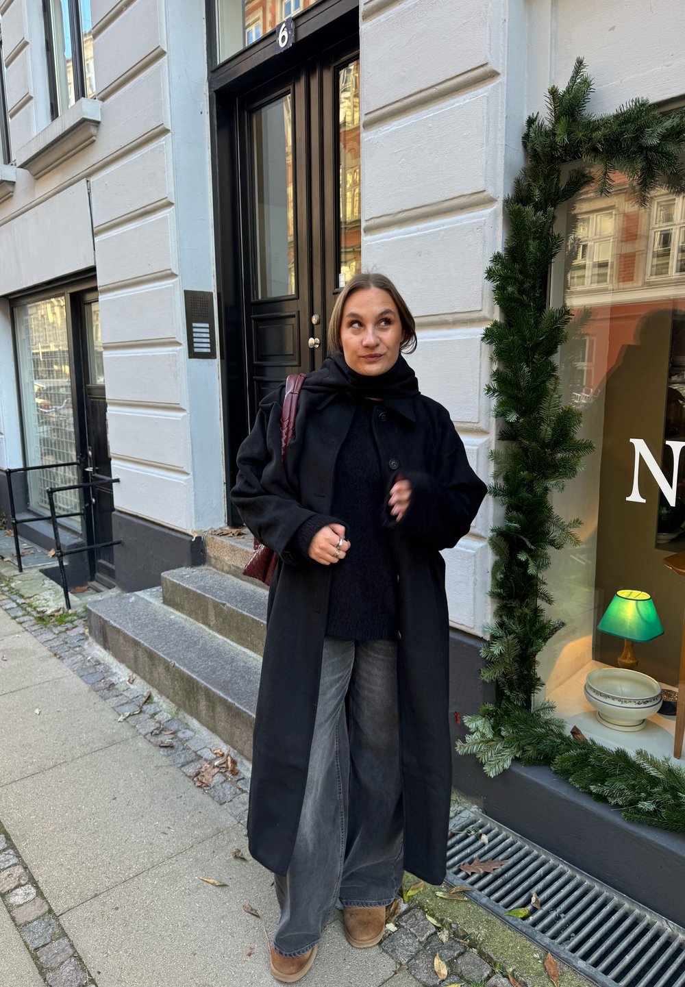 Black coat over a dark knitted jumper, grey wide-leg jeans, and brown boots. A burgundy bag and pavement with greenery and steps visible.