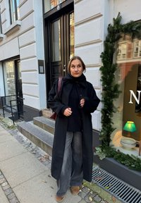 Black coat over a dark knitted jumper, grey wide-leg jeans, and brown boots. A burgundy bag and pavement with greenery and steps visible.