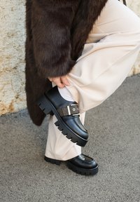 Zwarte leren schoenen met een stevige zool en een gestructureerde bruine band met een metalen gesp. Gecombineerd met lichtgekleurde broek.