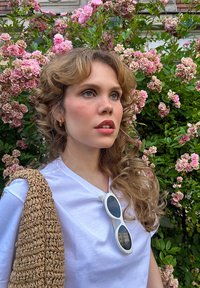 T-shirt blanc, sac tissé marron clair, lunettes de soleil blanches surdimensionnées, cheveux ondulés marron clair, entouré de fleurs roses et de feuillage vert.