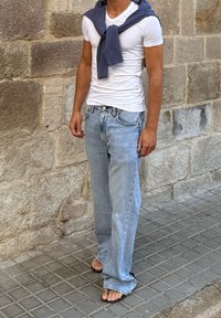 Bela prilegajoča majica, ohlapni svetlo modri jeans s raztrganim robom, temno moder pleten pulover, ki je premetan okoli vratu, črne japanke, stoječe na kamniti ploščadi.