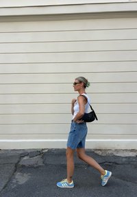 Mulher a caminhar de calções de ganga e uma t-shirt branca, a usar óculos de sol pretos e sapatilhas azuis e amarelas, a carregar uma mala preta.