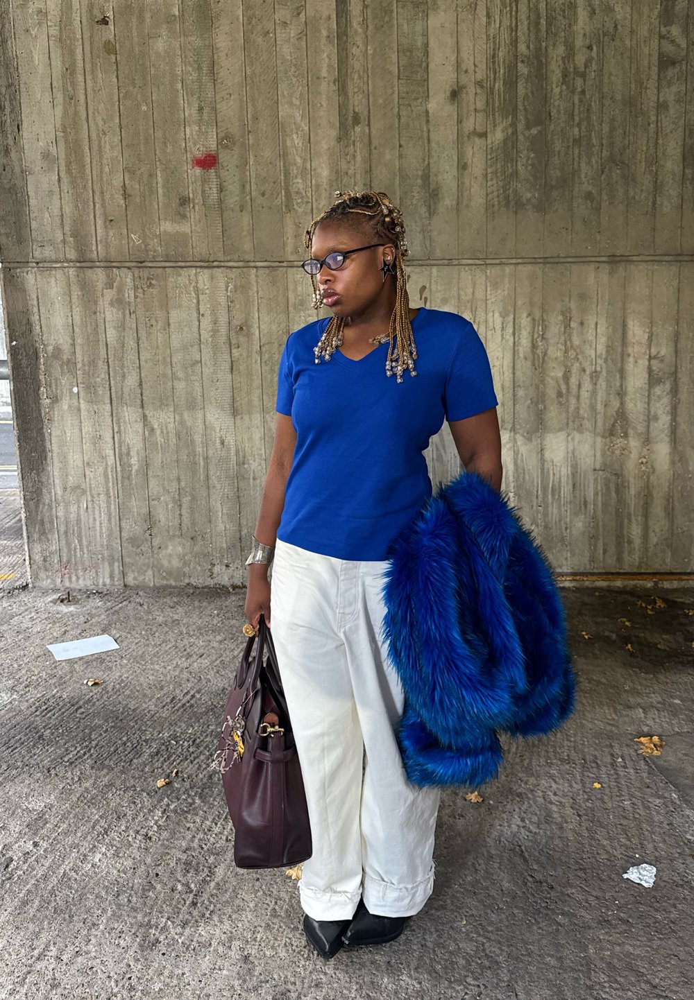 Blaues T-Shirt mit kurzen Ärmeln und V-Ausschnitt, weiße Hosen mit weitem Bein, dunkle Handtasche und ein blauer Kunstpelzmantel, mit Perlenakzenten im geflochtenen Haar.