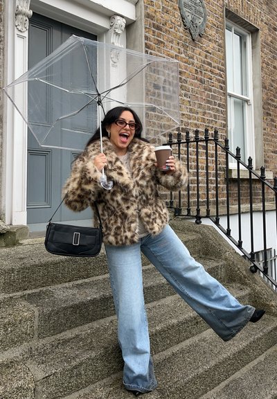 Paraguas transparente sostenido sobre una persona con un abrigo de piel sintética color beige, un bolso marrón con estampado, jeans acampanados de color azul claro y gafas de sol, en unas escaleras.