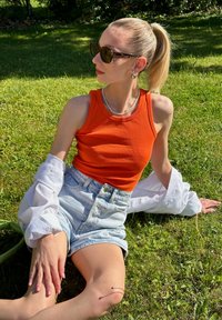 Camisola de alças laranja com nervuras, calções de ganga claros, camisa de manga longa branca colocada por cima. Acess�órios incluem óculos de sol e brincos pendentes.