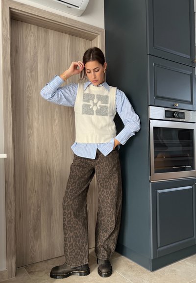 Chaleco de punto blanco con diseño gris, superpuesto a una camisa de rayas azul. Pantalones de pierna ancha con estampado de leopardo marrón y botas de tobillo negras. Fondo de cocina moderna.