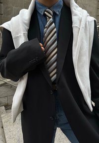 Μαύρο σακάκι με σχέδιο πάνω από πουκάμισο τζιν, ριγέ γραβάτα σε καφέ, μπλε και λευκά χρώματα, με ένα λευκό φούτερ ριγμένο στον ώμο.