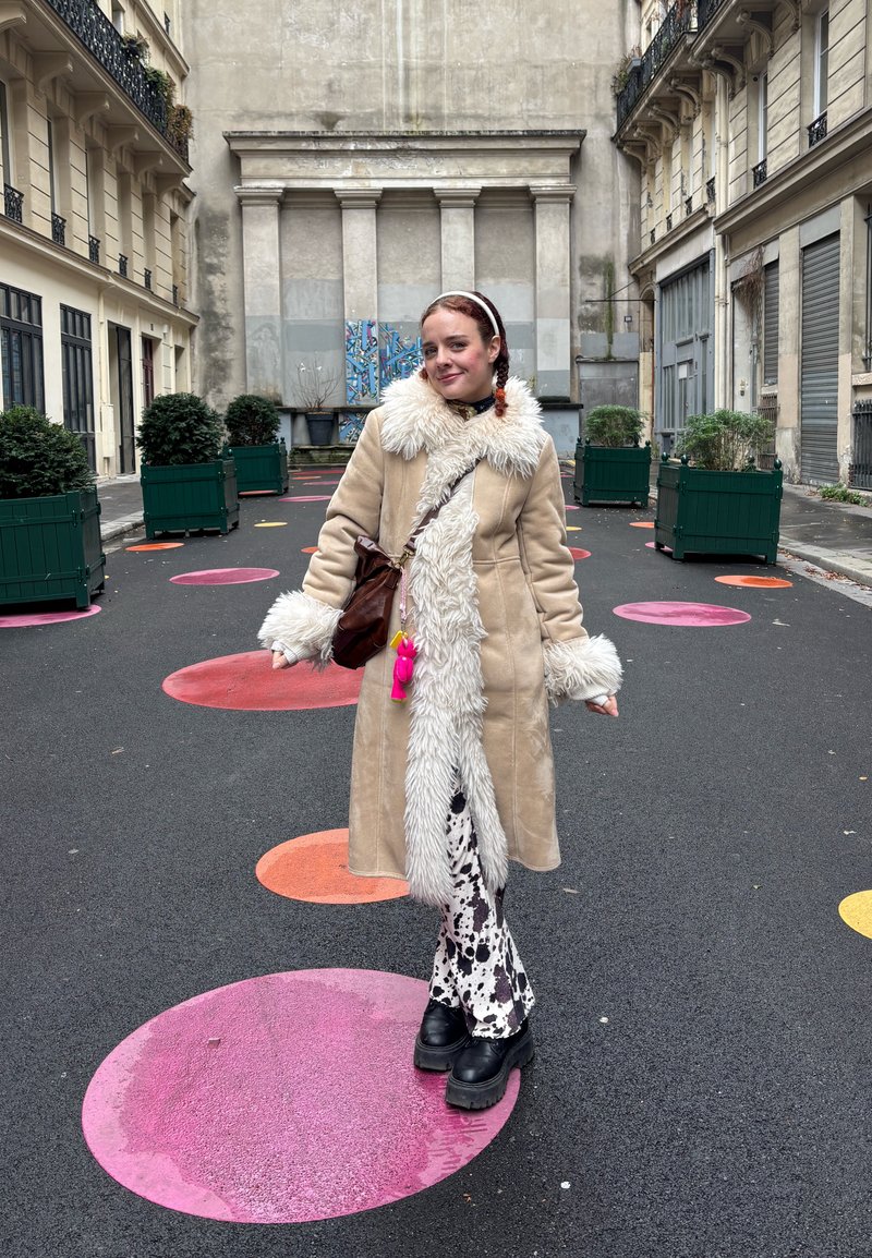 Cappotto lungo beige con collo e polsini in pelliccia sintetica, pantaloni neri svasati con motivo e stivali neri massicci, su uno sfondo di pavimentazione circolare colorata.