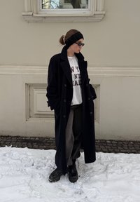 Long manteau en laine noir sur un sweat-shirt gris avec texte, pantalon gris ample et bottes noires épaisses. Sol couvert de neige et mur neutre en arrière-plan.