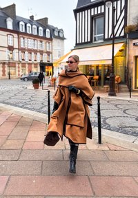 Poncho marron en laine avec un design drapé et des bordures contrastantes. Porté sur une tenue noire, associé à des bottes noires montantes et des gants.