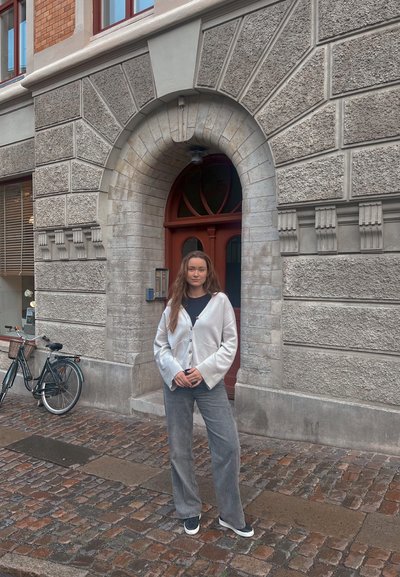 Cardigan plateado con botones, top negro, pantalones anchos grises y zapatillas negras, de pie frente a un edificio de piedra y una bicicleta.
