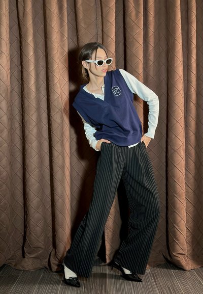 Top sin mangas azul marino sobre una camisa blanca de manga larga, combinado con pantalones de pierna ancha a rayas blancas y negras y gafas de sol blancas. Fondo texturizado.