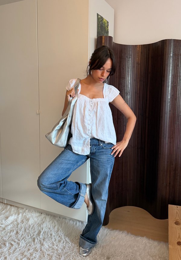 Femme debout à l'intérieur sur un tapis blanc, portant un chemisier blanc, un jean bleu, des chaussures argentées et tenant un sac à main argenté sur l'épaule.