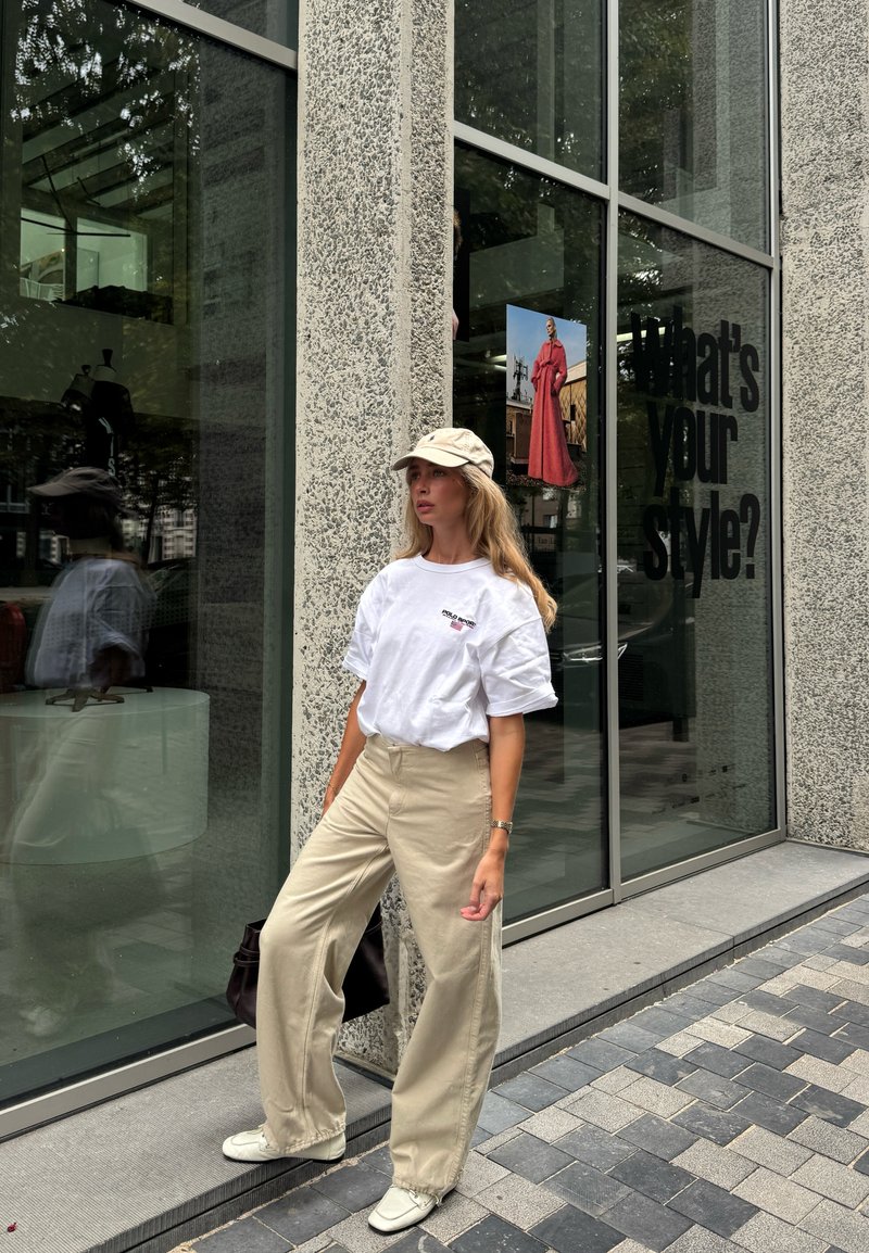Donna che indossa pantaloni beige, t-shirt bianca oversize, cappellino e scarpe bianche posa accanto a un edificio con il testo "What's your style?" sulla finestra.
