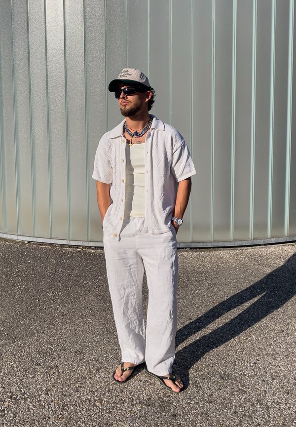 Mann trägt weiße Kleidung, Sonnenbrille, Kappe und Flip-Flops, steht mit den Händen in den Taschen auf Kies vor einer gebogenen Metallwand.