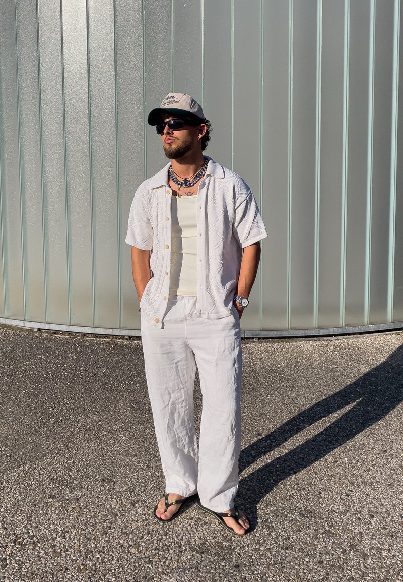 Homme portant une tenue blanche, des lunettes de soleil, une casquette et des tongs, debout sur du gravier, les mains dans les poches, devant un mur métallique courbé.