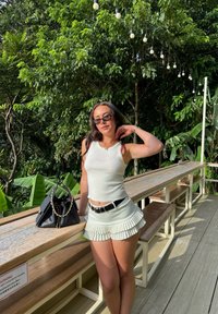 Vaaleanvihreä tankkitoppi ja siihen sopiva laskostettu hame mustalla vyöllä. Musta olkalaukku ketjusomisteilla. Ympärillä rehevää vihreyttä ja puisia huonekaluja.