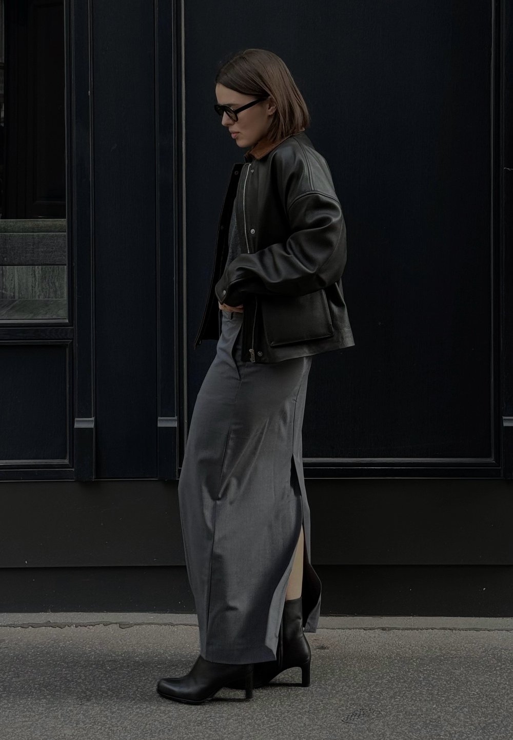 Melnas ādas bomberjakas nēsāšana virs pelēka maxi svārkiem ar sānu šķēlumiem. Melnas papēžu zābakus un tumšas brilles papildina izskatu uz tumša fona.