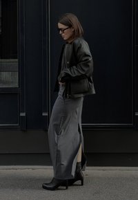 Melnas ādas bomberjakas nēsāšana virs pelēka maxi svārkiem ar sānu šķēlumiem. Melnas papēžu zābakus un tumšas brilles papildina izskatu uz tumša fona.