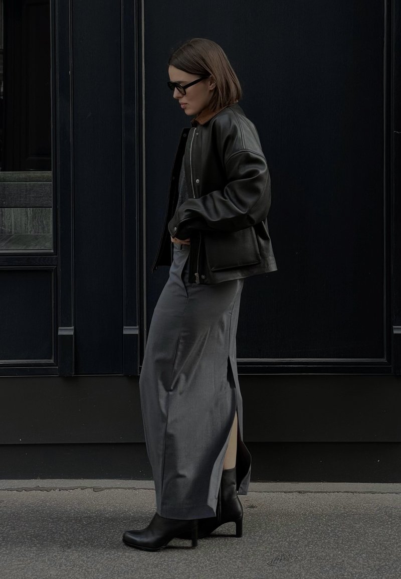 Μαύρη δερμάτινη bomber jacket πάνω από μια γκρι μακριά φούστα με πλαϊνές σχισμές. Μαύρες μπότες με τακούνι και σκούρα γυαλιά ολοκληρώνουν το σύνολο σε έναν σκοτεινό φόντο.