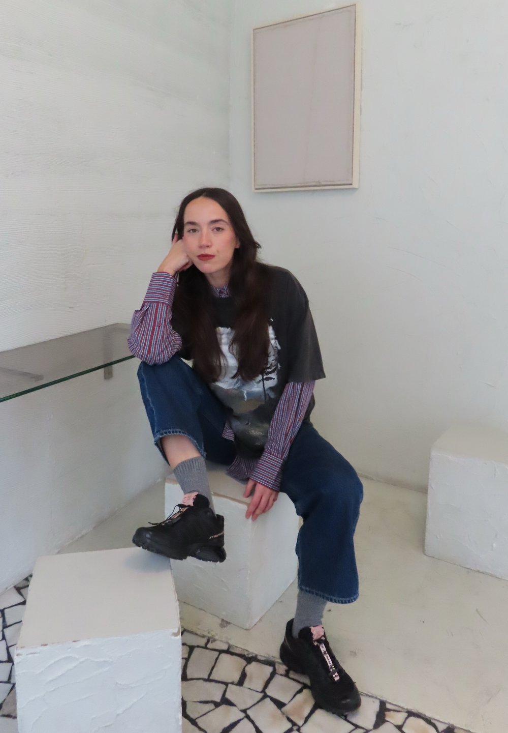 Sorte sneakers, grå crew-sokker, løstsiddende blå jeans og en sort grafisk t-shirt lagdelt over en stribet langærmet trøje. Hvid tekstureret baggrund.