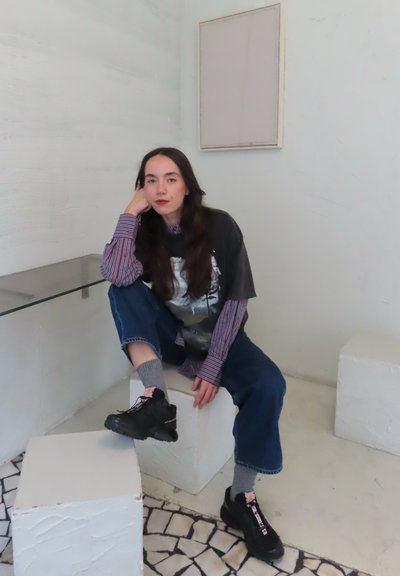 Zapatillas negras, calcetines grises hasta la mitad de la pantorrilla, vaqueros azules holgados y una camiseta negra con un gráfico encima de una camisa de manga larga a rayas. Fondo blanco texturizado.