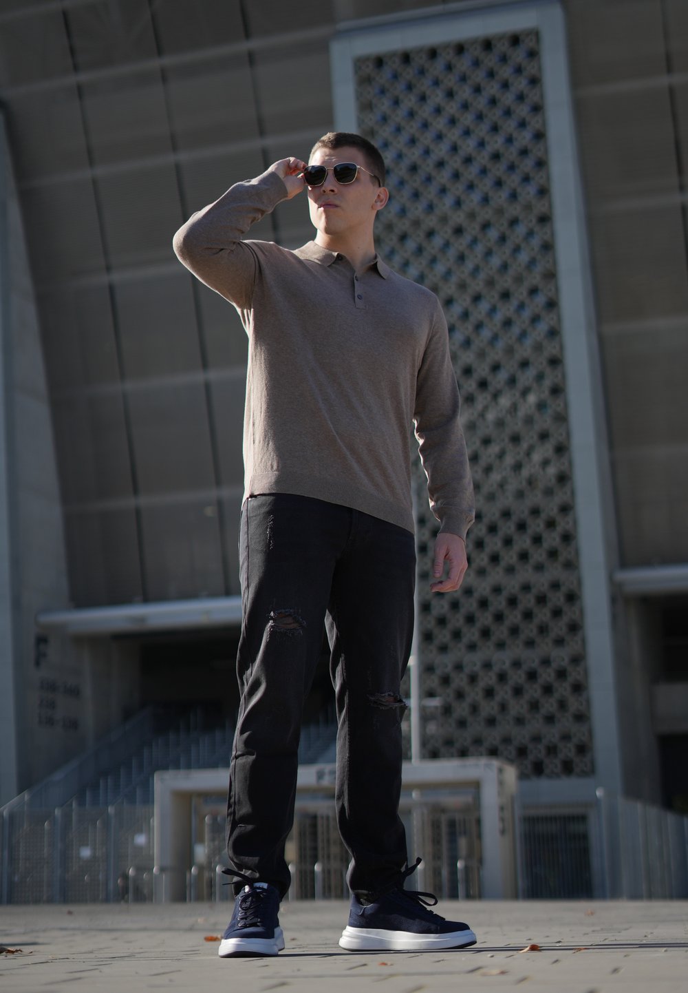 Light brown long-sleeve polo shirt, black distressed jeans, and navy sports shoes. Sunglasses reflect light; modern architectural background.