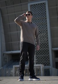 Light brown long-sleeve polo shirt, black distressed jeans, and navy sports shoes. Sunglasses reflect light; modern architectural background.