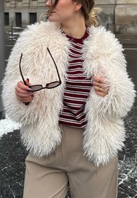 Personne portant une veste blanche duveteuse et une chemise rayée bordeaux, tenant des lunettes de soleil, debout sur un trottoir urbain enneigé.